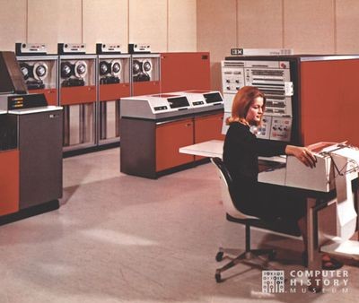 Eine Frau sitzt an der Operatorkonsole eines IBM /360 Systems; im Hintergrund links Kartenleseeinheit, 2 Diskdrives und 4 Tapestationen, alles in der typisch roten Farbe der 360er Reihe.
